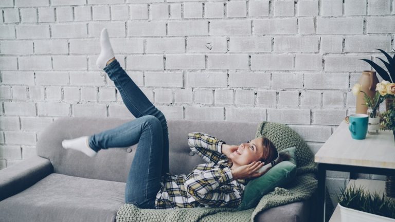 Joyful young woman is singing, listening to music in headphones and moving hands and legs dancing lying on sofa in modern loft style apartment. People, technology and fun concept.
